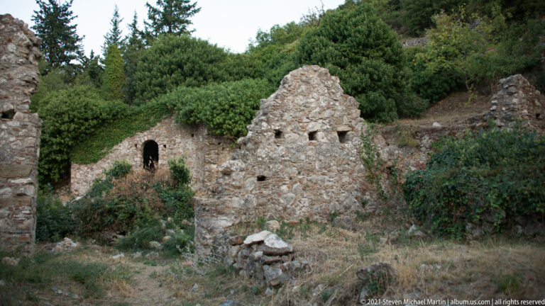 Small roofless gabled stone ruin at Mystras surrounded by shrubs, with another masonry opening partly visible behind.