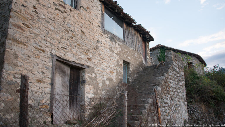 Stone hillside building at Mystras with wooden door, small window, timber upper section, tiled roof, and adjacent masonry stair.