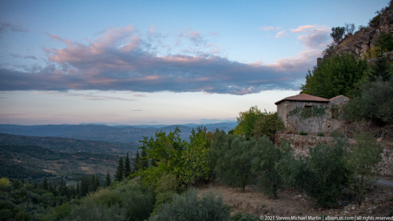 Stone building and olive-covered hillside at Mystras overlooking a broad valley beneath evening clouds.
