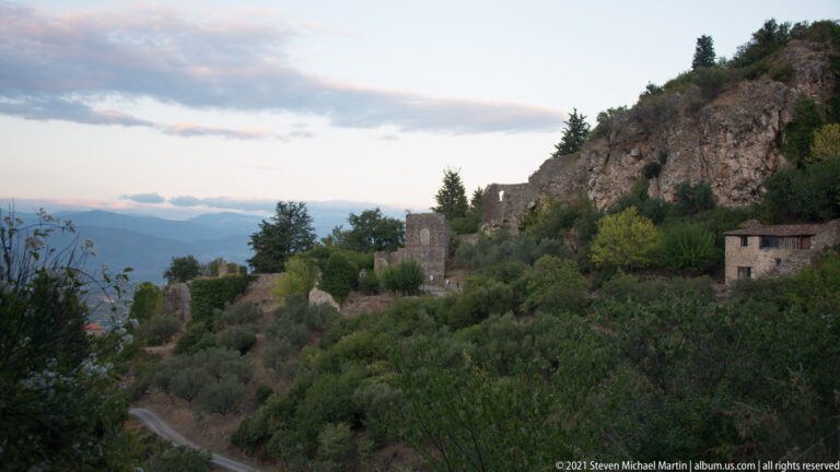 Scattered stone ruins and buildings on the wooded Mystras hillside with a winding path and distant mountains.