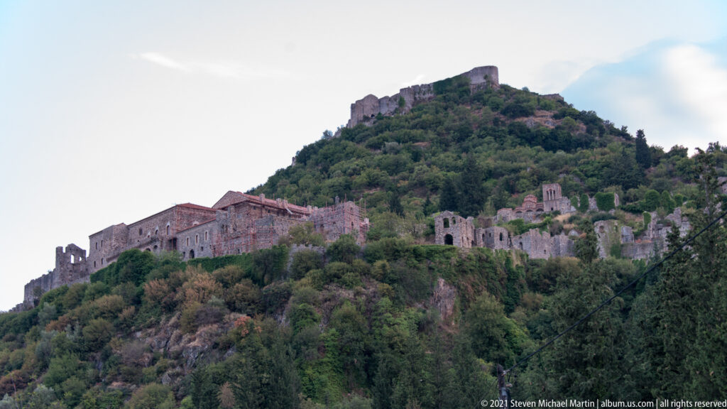 Panoramic view of Mystras with large medieval complexes, church ruins, and summit fortifications rising above the wooded hillside.