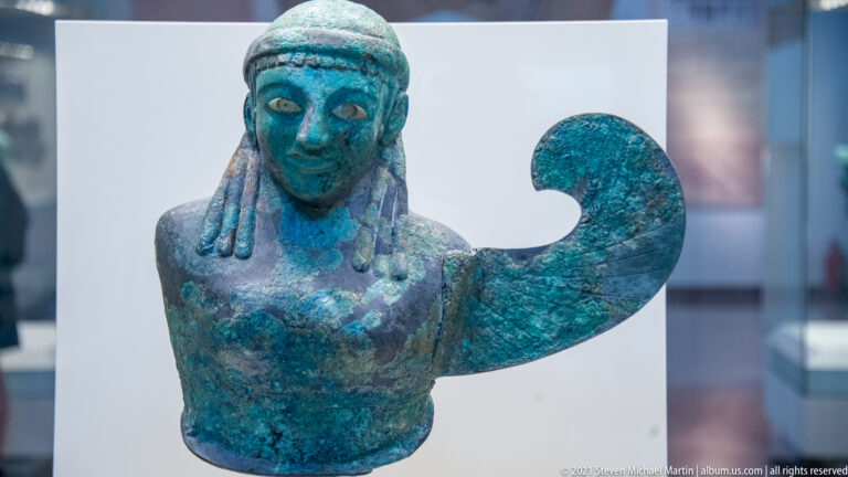 Blue-green bronze bust with stylized face, long hair, and one large curved raised arm.