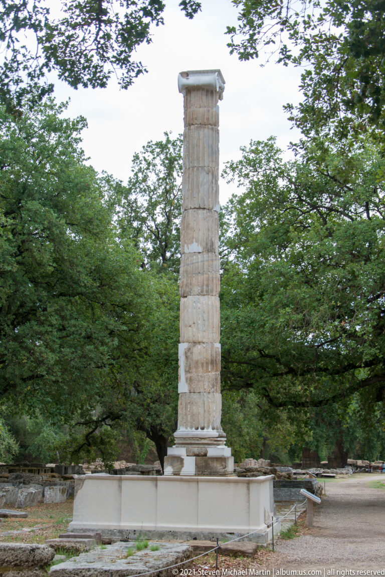Single restored fluted stone column standing on a low ancient foundation amid trees.