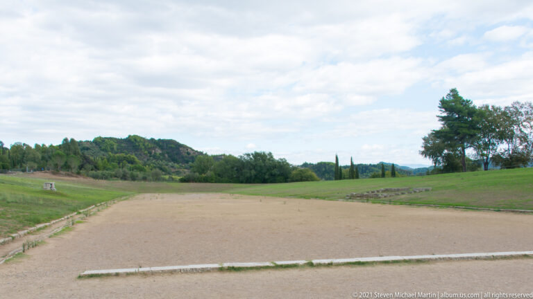 Broad earthen running track of the ancient Stadium of Olympia between long grassy spectator embankments.