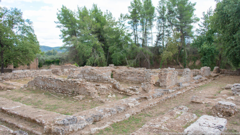 Low brick and stone walls forming rectangular rooms among pine trees at Ancient Olympia.