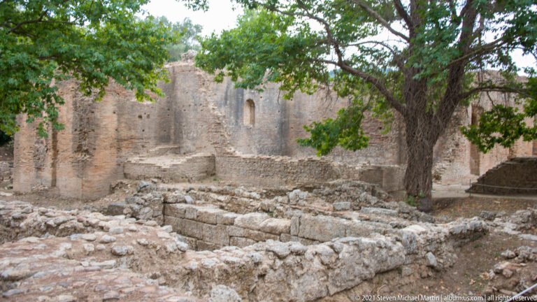 High ancient brick walls and low stone foundations beneath a large tree at Ancient Olympia.