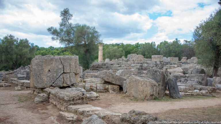 Large fallen stone blocks and broken architectural fragments scattered among low foundations, with a distant standing column.