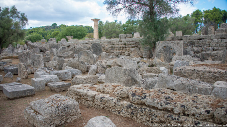 Dense field of weathered fallen stone blocks and architectural fragments with a standing column in the distance.