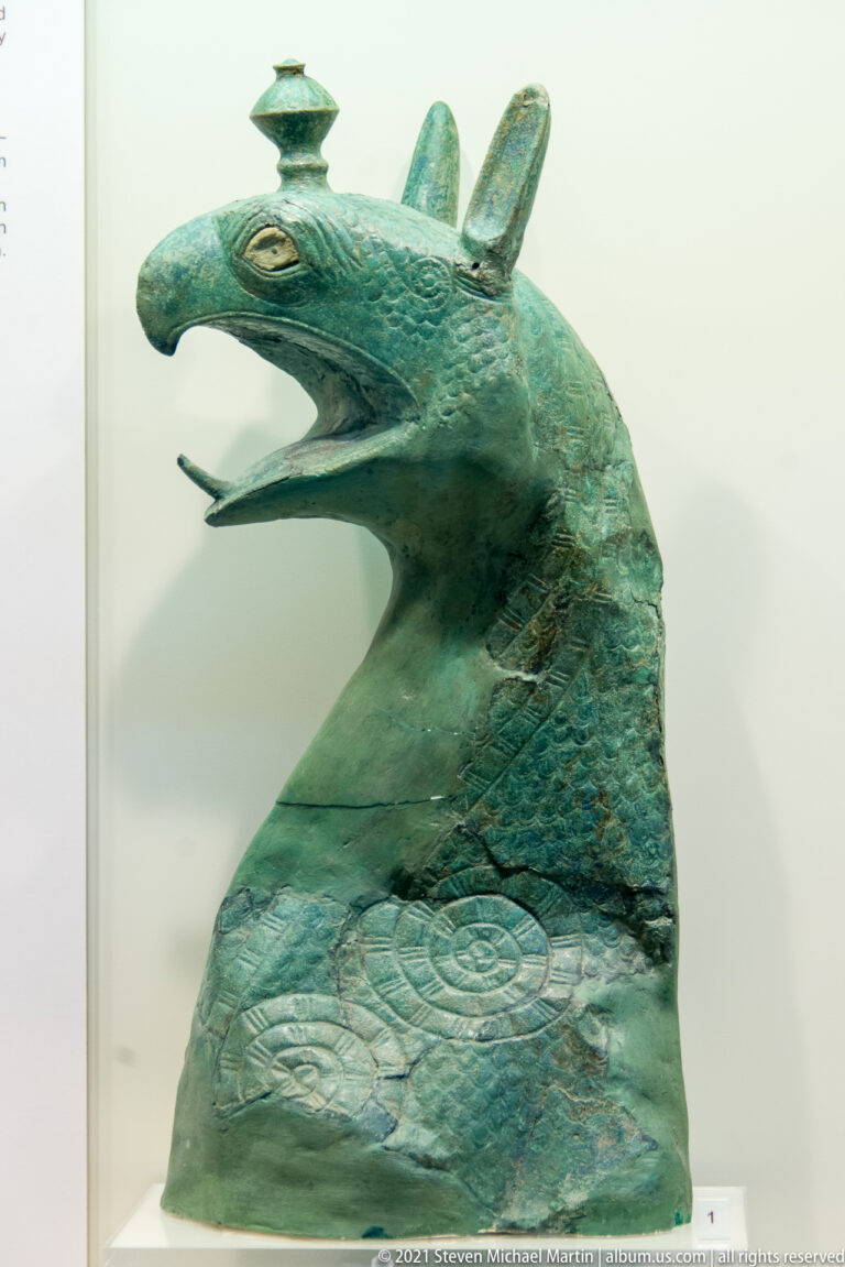 Archaic bronze griffin-head cauldron protome from Olympia, with hooked open beak, pointed ears, long neck, and incised decoration.