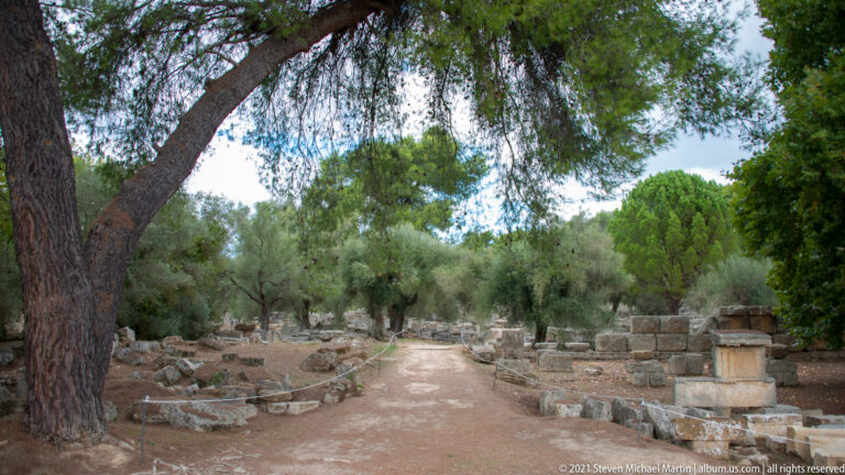 Tree-lined earthen path running between low ruins and scattered stone blocks at Ancient Olympia.