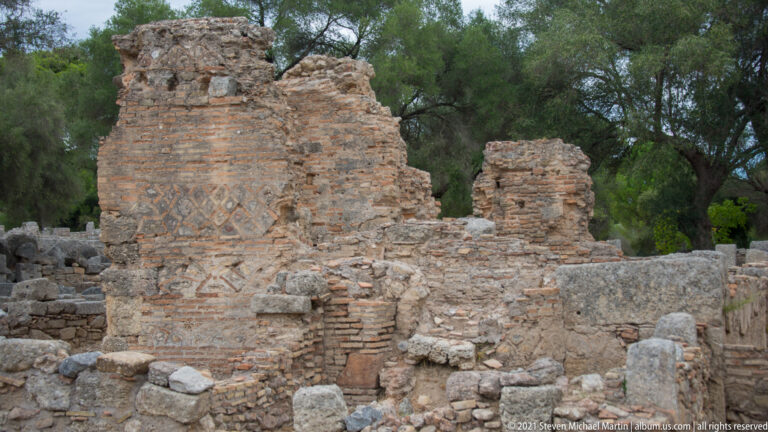 Tall ruined wall with horizontal brick courses and geometric diagonal brick patterns amid stone rubble.