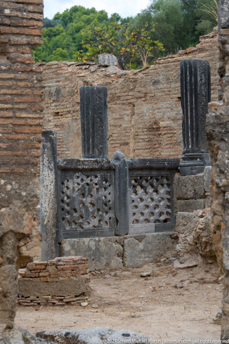Low pierced marble screen between dark fluted columns and masonry piers inside ancient brick ruins.