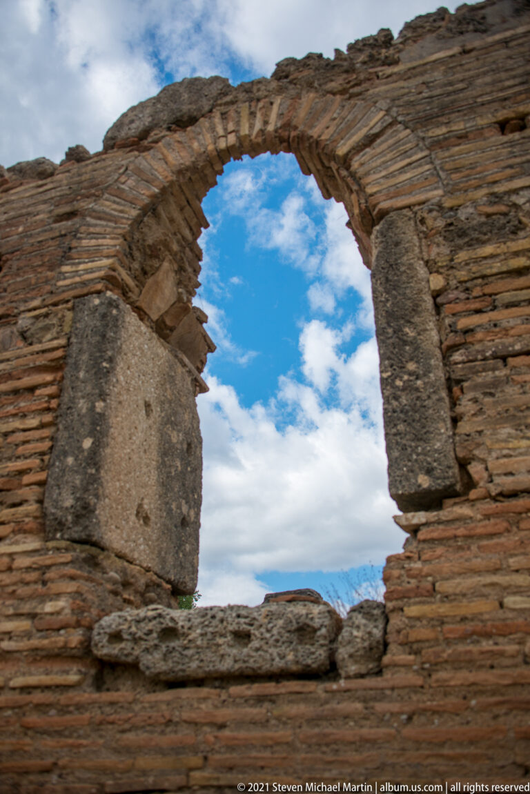 Round brick arch framed by large stone blocks in a ruined wall, opening onto blue sky.