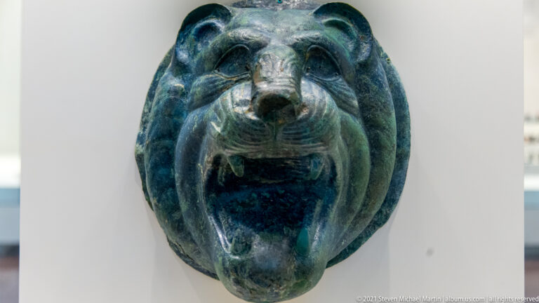 Frontal dark-green bronze lion head with rounded ears, open mouth, teeth, and deeply modeled muzzle.