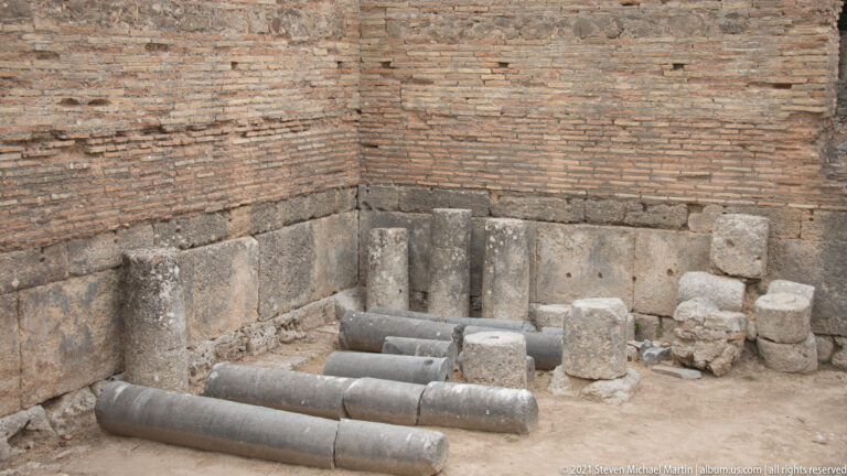 Several dark stone column shafts lying on the floor beside short bases inside ancient brick walls.