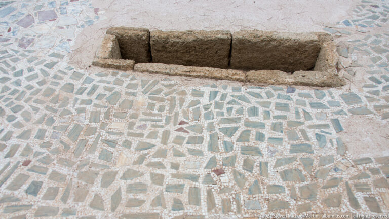 Geometric pale mosaic floor surrounding a long rectangular stone-lined opening.