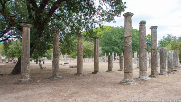 Rows of slender ancient stone columns surrounding an open tree-shaded courtyard at Ancient Olympia.