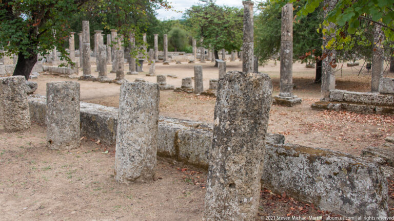 Rows of weathered stone columns and low walls surrounding an open tree-shaded court at Ancient Olympia.