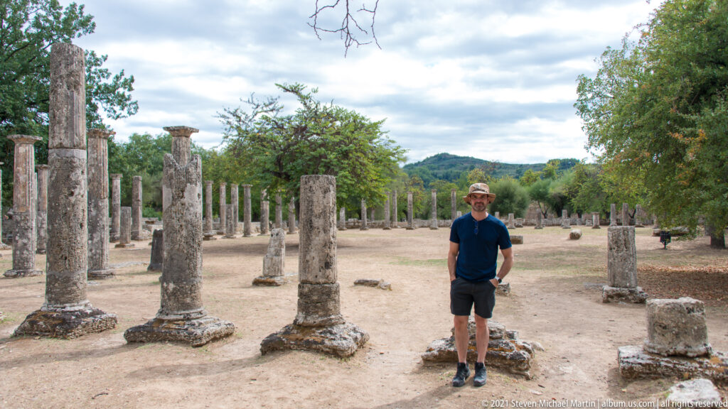 Man standing among rows of ancient stone columns and bases in an open courtyard at Olympia.
