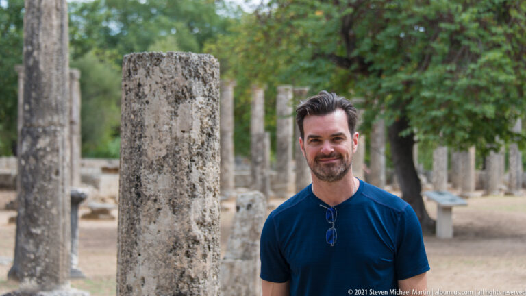 Close portrait of a man beside a weathered ancient column, with more columns blurred behind.