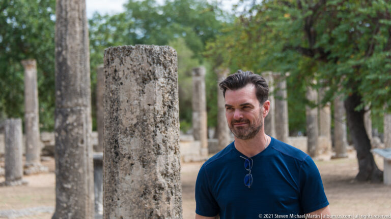 Three-quarter portrait of a man beside a broad stone column with rows of ancient columns behind.