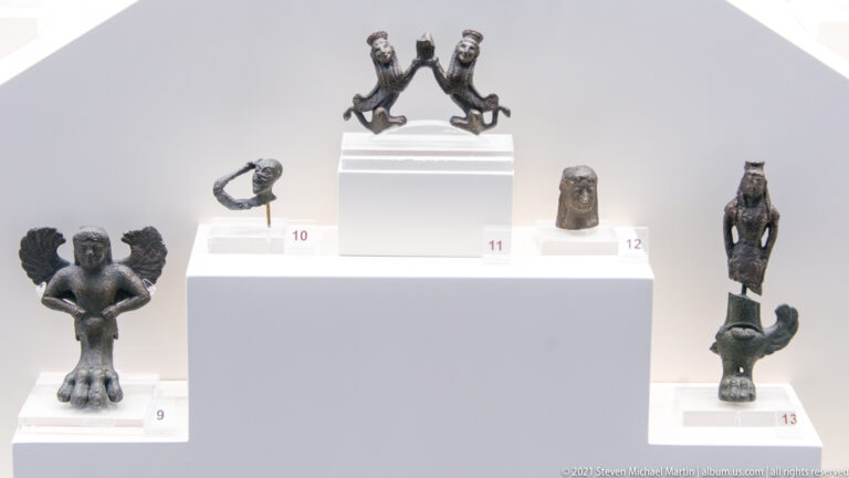 Several small ancient bronze human and animal figurines displayed on stepped white museum mounts.