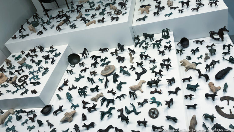 Large museum display filled with hundreds of small bronze animal and human figurines on stepped white platforms.