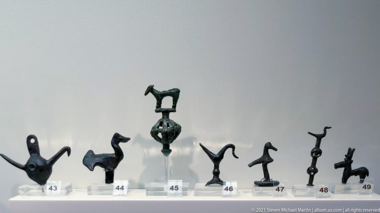 Row of small bronze animal and geometric votive figures on individual museum stands with numbered labels.