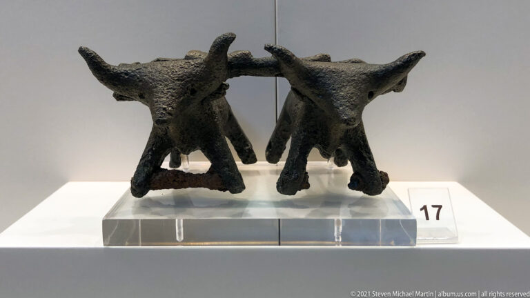 Two small dark bronze horned animal figures displayed side by side on a clear stand.