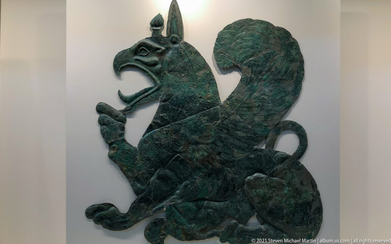 Large green-patinated bronze griffin in profile with open beak, raised foreleg, and broad curled wing.