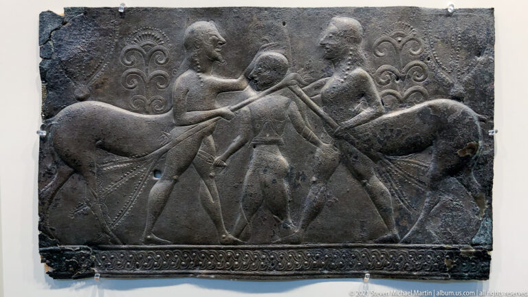 Bronze relief with two bearded centaurs facing a smaller central human figure amid scrolling ornament.
