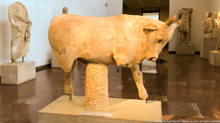 Large ancient bull sculpture with short horns and warm reddish surface coloration displayed in a museum gallery.