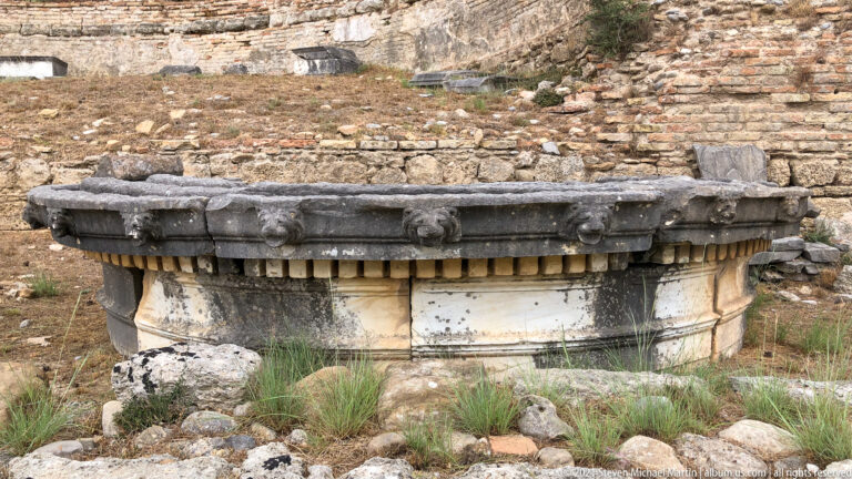 Low circular stone structure with projecting carved lion heads and a dentil-like decorative band.