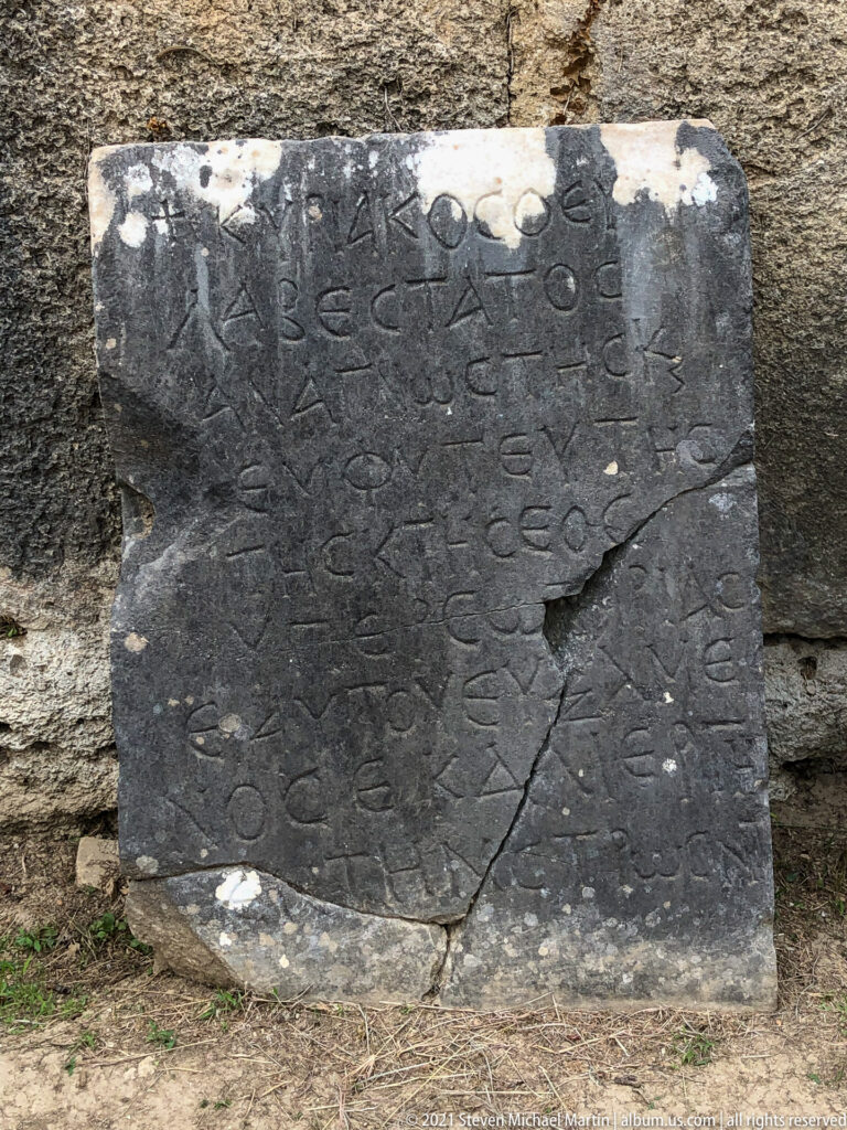 Large dark stone slab with several lines of ancient Greek inscription, cracks, and pale surface losses.