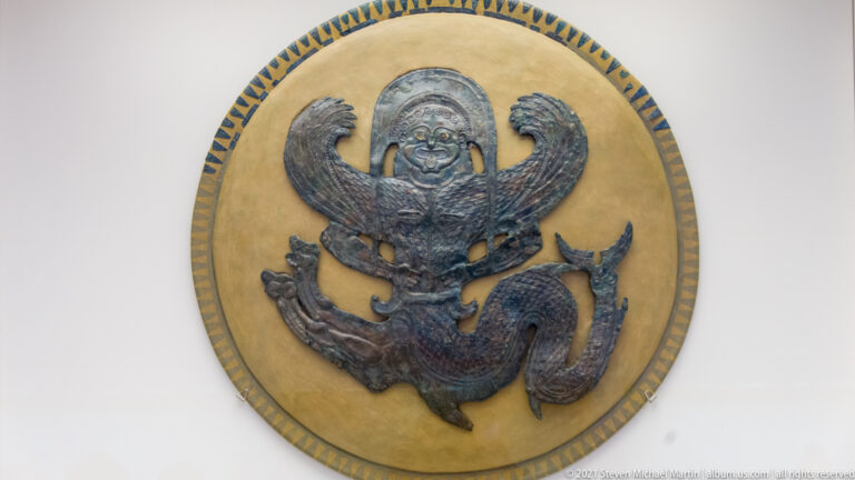 Dark bronze winged mythological figure mounted on a round tan backing with a patterned border.