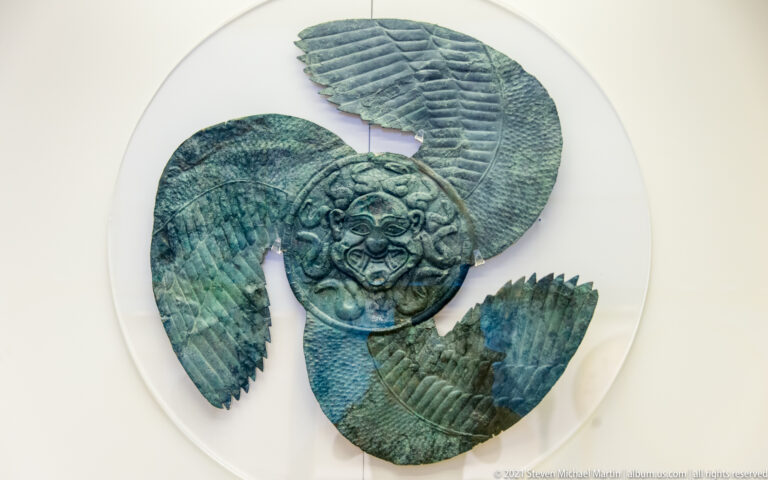 Green bronze ornament with three radiating feathered wings around a circular mask-like face.