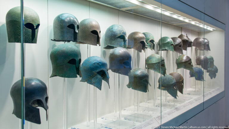 Rows of ancient Greek bronze helmets in the Archaeological Museum of Olympia, including enclosed and open forms with varied cheek pieces.