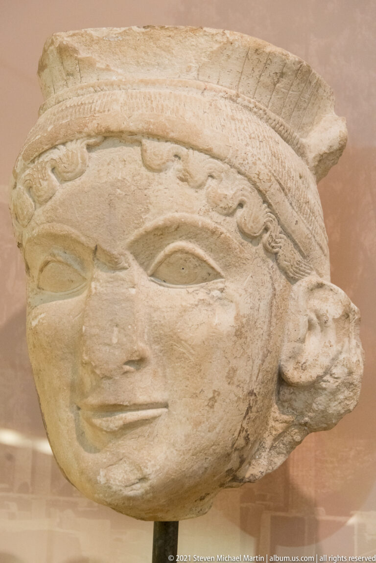 Large pale terracotta head with almond eyes, archaic smile, patterned headdress, and broken edges.