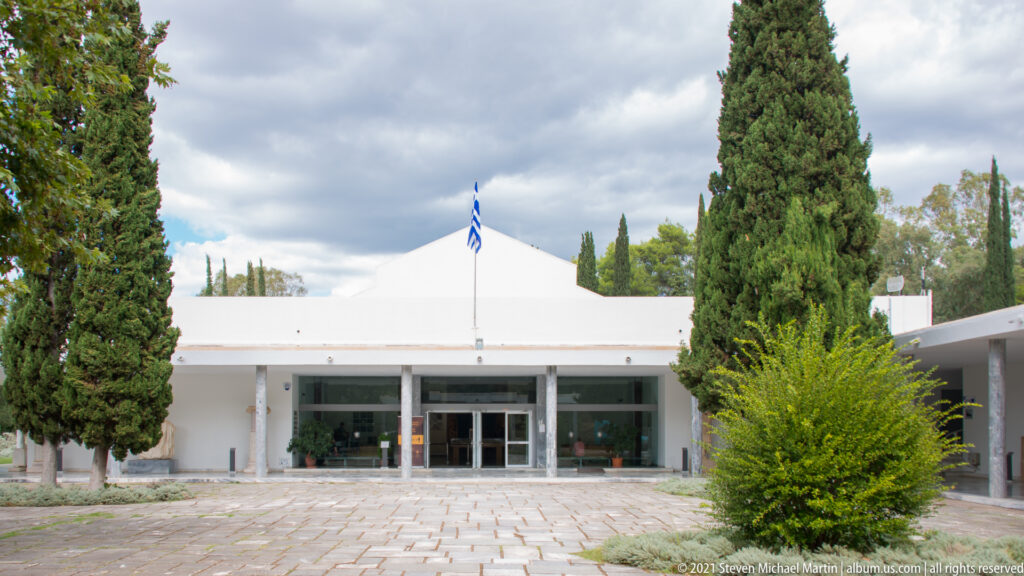 White museum entrance with Greek flag, glass doors, paved forecourt, and tall cypress trees.