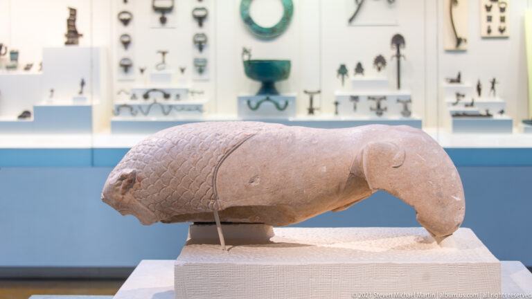 Profile of a fragmentary pale stone animal with carved scales on its front body and broken extremities.