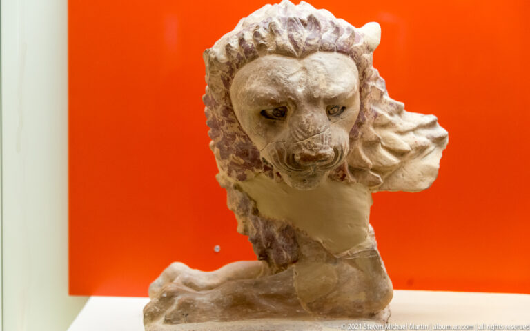 Fragmentary pale marble lion with deeply carved mane, broad muzzle, and broken body against a red background.