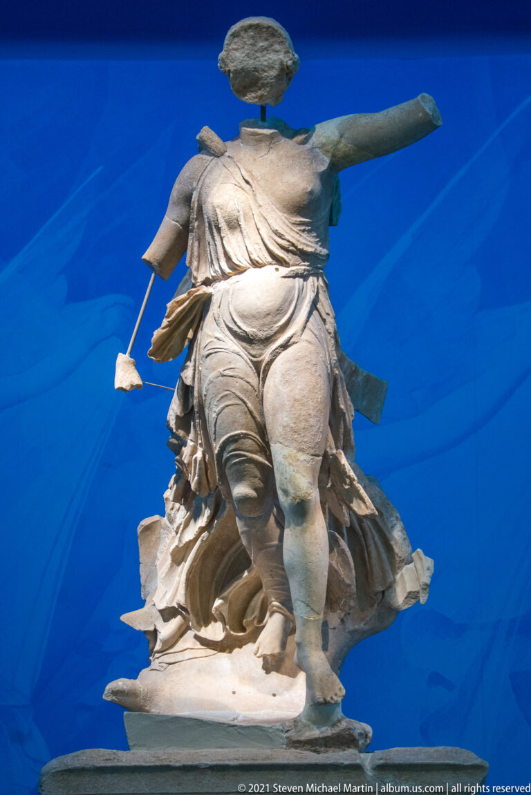Fragmentary pale stone female figure with flowing drapery and one arm extended sideways against a blue background.