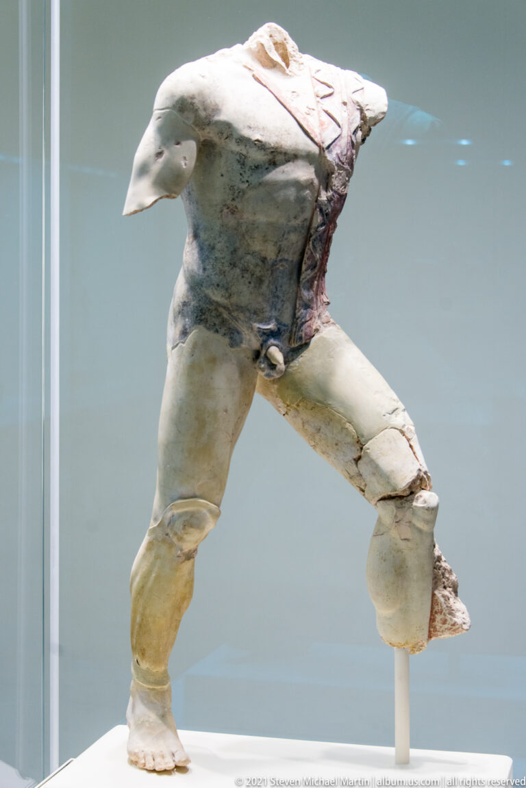 Headless and armless marble male figure in a wide lunging stance with muscular legs and twisting torso.