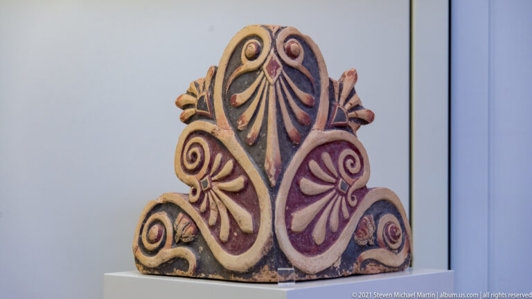 Painted triangular terracotta roof ornament with central palmette, curling volutes, and red, cream, and dark colors.