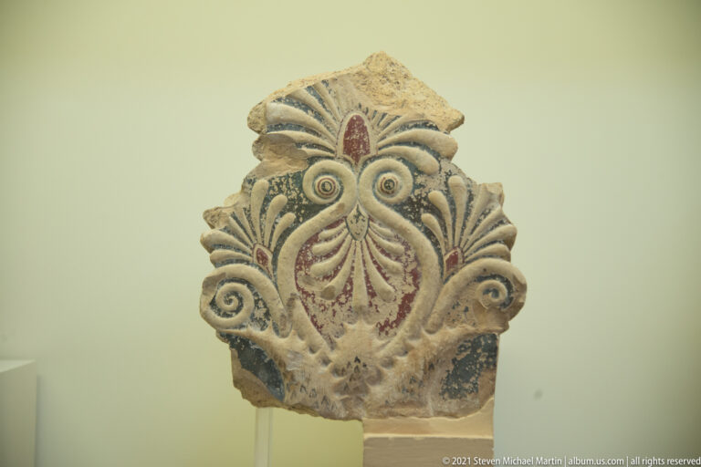 Painted terracotta architectural ornament with a central palmette, curling volutes, layered leaves, and surviving red and blue-green pigment.