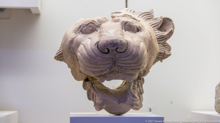 Frontal carved lion head with open mouth, rounded ears, broad muzzle, and radiating mane.