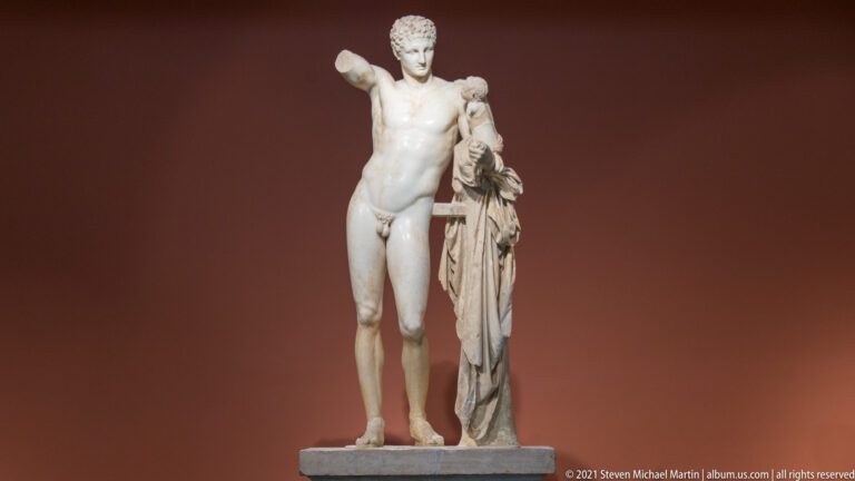 Full-length marble group of Hermes carrying infant Dionysus, with Hermes leaning beside a cloak-draped tree-trunk support.
