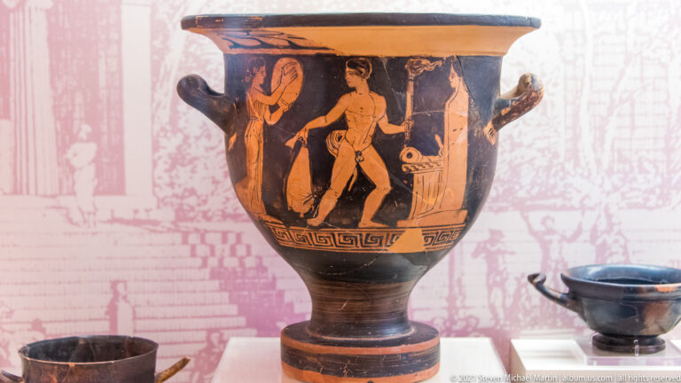 Large red-figure krater with black ground, tall handles, and painted male figures around the vessel body.