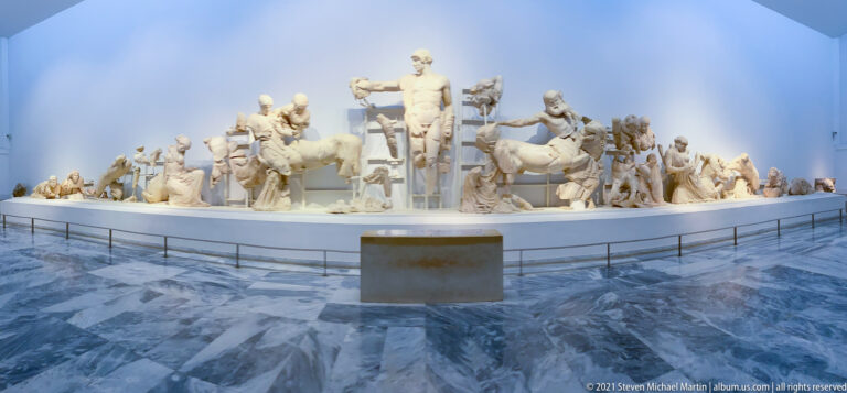 Oblique gallery view of fragmentary marble figures and horses arranged as a monumental Greek temple pediment.