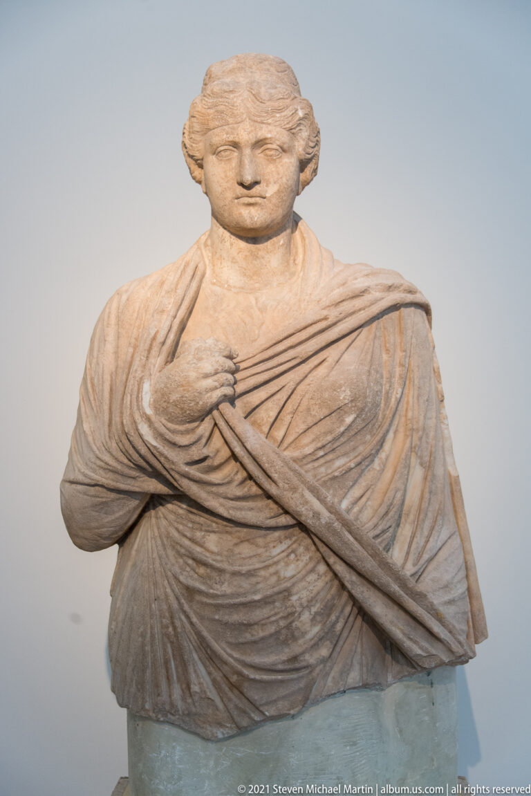 Stone female portrait bust with center-parted hair, layered drapery, and one hand resting against the chest.